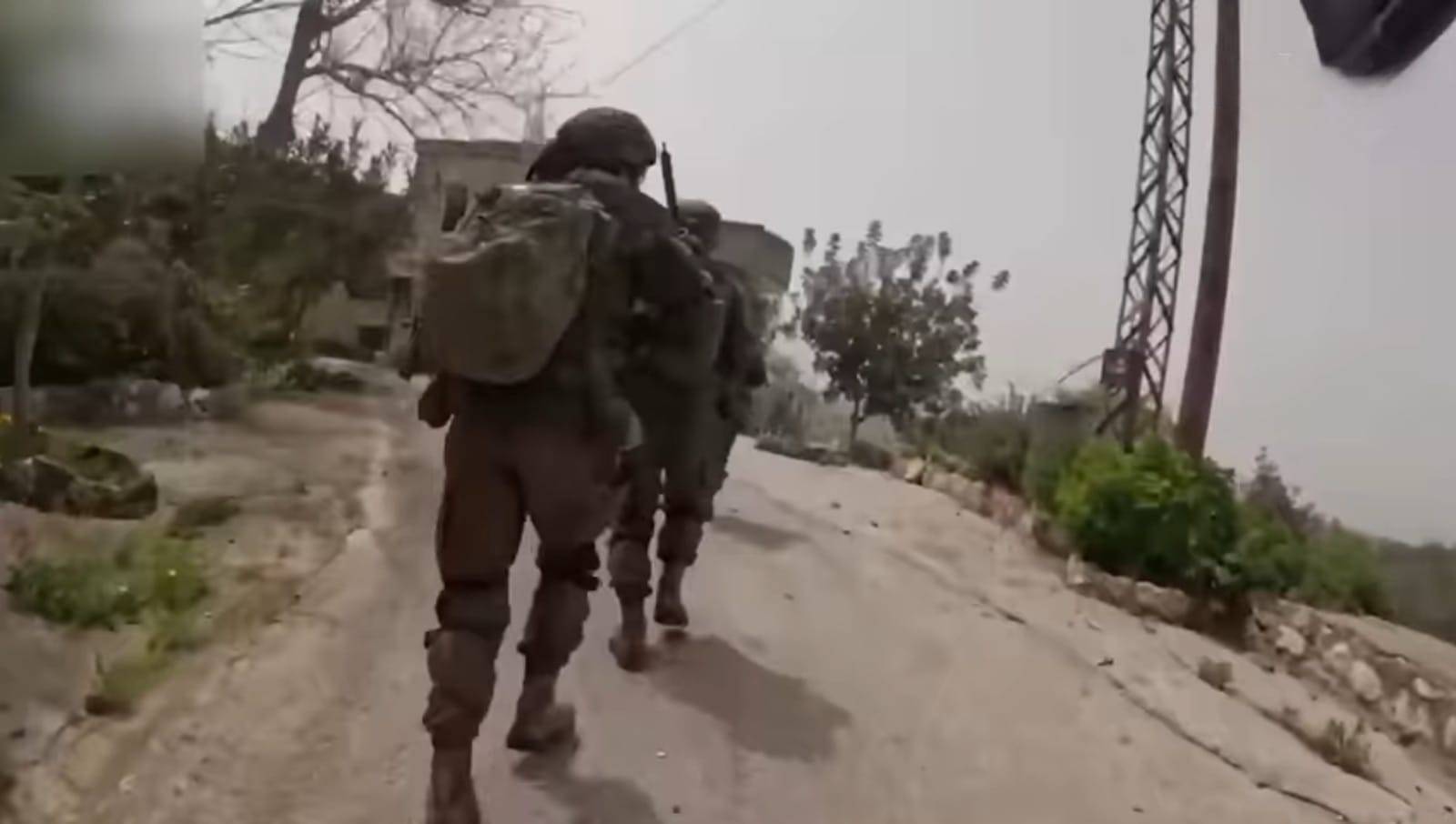 Two soldiers are running toward a house along a road lined with bushes and trees on both sides.