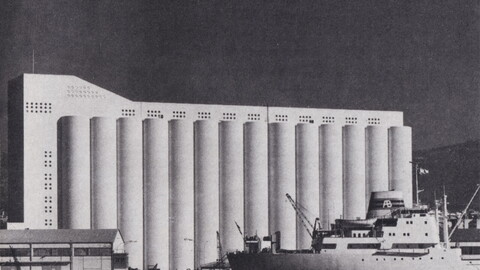 A black and white image of the Beirut grain silos which consist of a white concrete building with 14 cylinder shapes. The sea and a boat can be seen in the foreground.