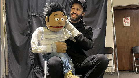 A smiling young man dressed in black is sitting on a chair and holding up a puppet.