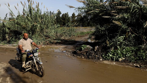 A man rides a motorcycle on a road filled with sewage water. Bamboo sticks protrude from the side of the road. 