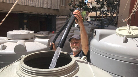 A man in a cap with a white mustache points a thick black hose into a plastic grey water container, and water flows from the hose into the container. There are other such containers around him.