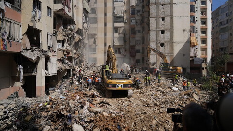 Two bulldozers and several people in high visibility vests stand atop a large mound of rubble, in the middle of a residential area, amid the destruction and ruin of nearby buildings.