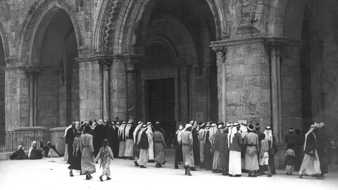 A black-and-white photograph of a group of men gathered outside the arched entrance of al-Aqsa Mosque in Jerusalem. 