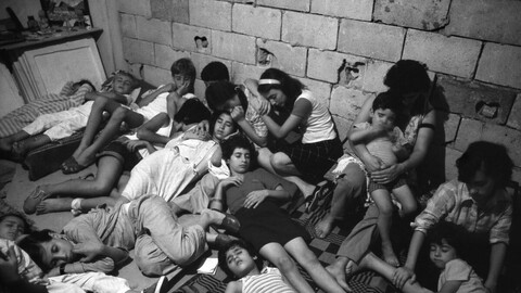 A black and white photo of a group of people leaning against one another, asleep on the floor, in what looks like a basement.