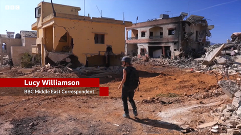 A woman stands amid rubble, center frame. Around her are fully demolished and semi-demolished buildings. She is wearing a helmet and flak jacket. To her left is a graphic that reads: “Lucy Williamson | BBC Middle East Correspondent”