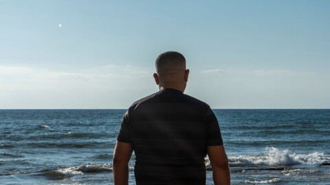 A man stands with his back to the camera and the sea before him. The sun is bright and the water is a deep blue. 