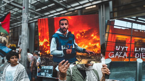 People hold up posters and signs at a protest. The poster in the center of the image is a photo of martyred Al Jazeera journalist Anas al-Sharif, killed by Israel on August 10, 2025. Next to it is a poster that reads: "NYT LIES, GAZA DIES"