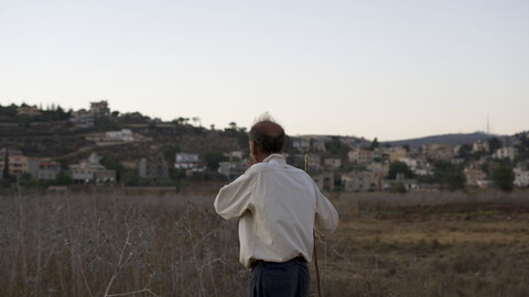 A man stands in a dry field looking toward a hillside town at dusk.