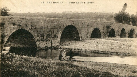 An arched stone bridge crosses the Beirut River; mountains are visible in the background. Three people are walking by the river. 