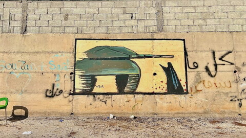 A mural of a green military tank is painted on a concrete wall with graffiti and a low brick barrier above; a woman is confronting the tank; trees and blue sky are visible in the background.