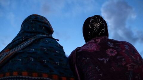 Two women wearing hijab stand under a clear blue sky.