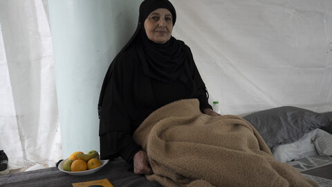 An older woman dressed in black sits beside a bed inside a tent-like shelter, holding a folded blanket. A small plate of fruit and a book rest on the bed next to her.