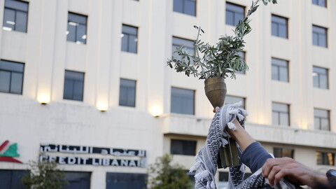 A hand holds remains of a missile filled with olive branches. A kuffiyeh is wrapped around the hand.