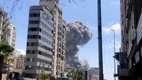 A dark grey plume of smoke rises behind a modern-looking building.