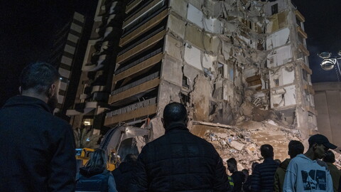 Crowd gathers at night before a partially collapsed building, with exposed interiors and debris. Somber scene with press and onlookers observing.