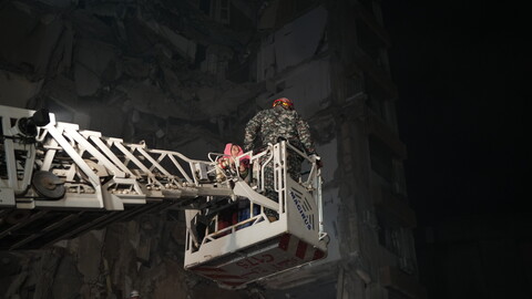 A nighttime photograph of a destroyed building. Rescue workers and first responders stand amid the rubble, dwarfed by the size of the building and its destruction. A lit-up crane reaches into the darkness and carries a survivor.