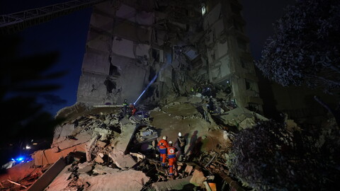 A nighttime photograph of a destroyed building. Rescue workers and first responders stand amid the rubble, dwarfed by the size of the building and its destruction.