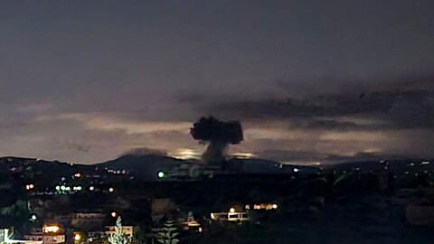 A dark plume of smoke rises into the night sky above a distant city, with lights from buildings visible in the foreground.
