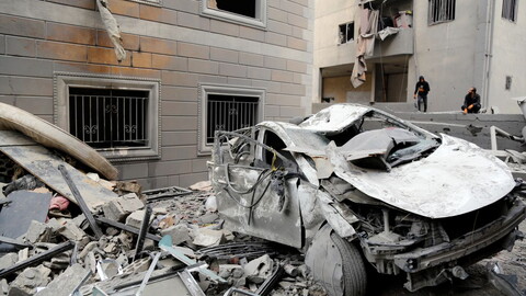 Heavily damaged car amidst rubble in front of a battered building. 