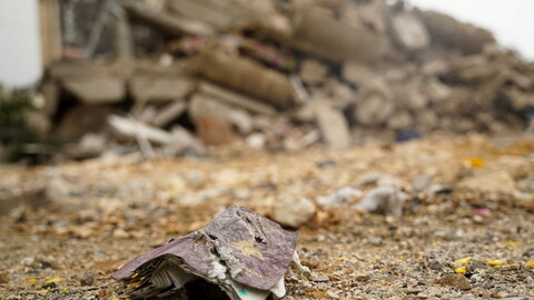 Tattered papers lie on rocky ground with a collapsed building in the background,