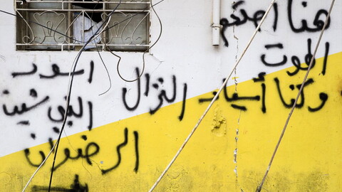Graffiti in Arabic covers a white and yellow exterior wall beneath a barred window with exposed wires hanging across the scene.