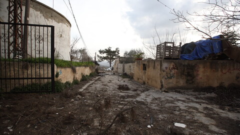 A narrow, muddy path between weathered concrete walls is flanked by a rusty gate and graffiti.