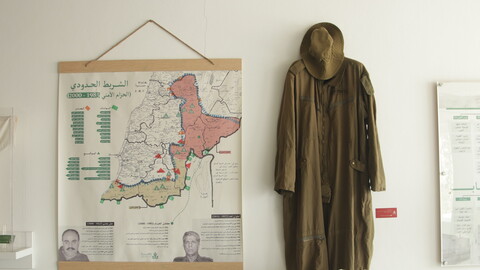 An exhibition wall featuring a large framed border map with Arabic text on the left, and a full-length olive-green military jumpsuit with a matching boonie hat hanging on the right.