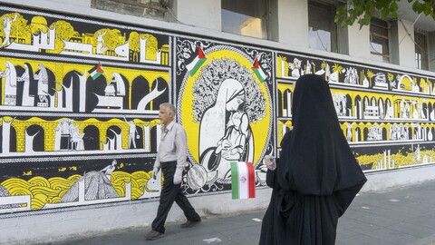 A side-view shot of a woman in holding a small Iranian flag as she walks along a city sidewalk. In the background, a man walks past a long, vibrant mural. The mural displays intricate illustrations, small Palestinian flags, and a central portrait of a mother cradling a child.