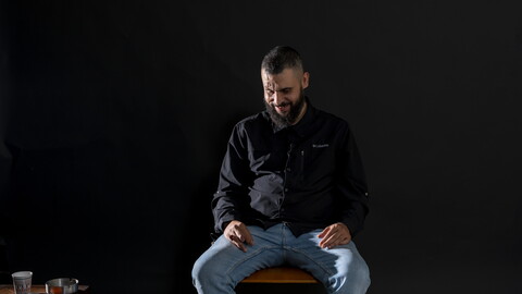 A man sits on a seat in front of a black background. He is bearded, wearing black, and looks down, laughing shyly.