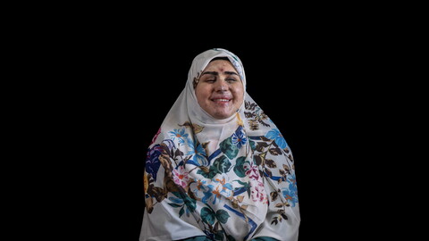 صورة استوديو بلقطة متوسطة لامرأة تُدعى روان الموسوي، تجلس أمام خلفية سوداء مصمتة. ترتدي حجاباً أبيض مزيناً بنقوش زهرية زاهية بألوان الأزرق والبنفسجي والبرتقالي، وفستاناً أخضر داكناً بأكمام طويلة. تبدو مبتسمة بابتسامة دافئة وعيناها مغمضتان، وتظهر على وجهها آثار ندوب وإصابات ناجمة عن شظايا في طور الالتئام، لا سيما في مناطق الجبهة والأنف والخدين.