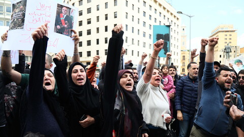 A crowd of people protest in a city street, raising their fists and shouting; one person holds a sign with Arabic writing and a photo of a man.