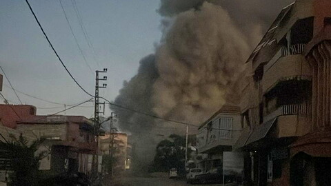 A street-level view in a residential neighborhood where a massive, towering plume of thick grey smoke rises high into the sky from directly behind the buildings on the right side of the road. A string of bare light bulbs hangs across the street under a dim sky.
