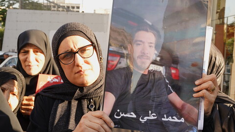 A woman in a black hijab and glasses holds up a large, laminated portrait of a missing man named Mohammad Ali Hassan during an outdoor demonstration. Other women stand closely behind her in solidarity.
