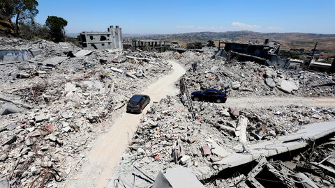 A high-angle panoramic view of massive structural destruction in Tebnine, South Lebanon. Mounds of shattered concrete and exposed rebar line a curved dirt road that has been cleared through the ruins. A dark gray hatchback and a black sedan are navigating the road amidst the completely flattened landscape.
