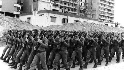 A black-and-white photograph of a large formation of Lebanese Army soldiers marching in tight unison during a drill. The soldiers are dressed in matching uniforms and combat boots, wearing distinctive helmets with integrated ear and chin protection, holding rifles diagonally across their chests. In the background, a war-torn urban landscape features large mounds of dirt and rubble in front of a heavily damaged, multi-story apartment complex and a smaller white building marked with bullet holes.