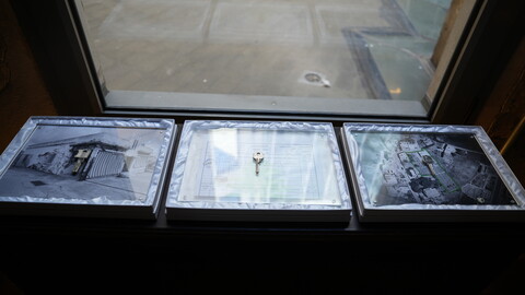 Three framed displays rest on a windowsill, each containing a key placed over a visual object: a photograph of a damaged house, a title deed, and an aerial image of a marked area. 