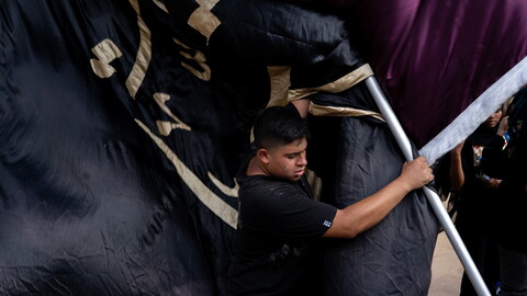 A young man wearing a black shirt strains as he hoists a large metal pole carrying heavy black and maroon religious banners trimmed with gold Arabic calligraphy during Ashoura commemorations.