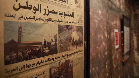 A framed Arabic newspaper with images of crowds, soldiers, and vehicles hangs on a textured wall next to a fire alarm.