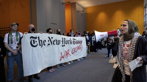 Protesters at a demonstration inside The New York Times building, holding a banner accusing the publication of coverage related to war crimes, with one person speaking through a megaphone.