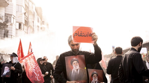 A man dressed in black walks through a crowd holding a framed portrait of Ayatollah Ali Khamenei against his chest while raising a red sign with the Arabic text "#انتقام" (#Revenge) in the air. Red flags and other mourners carrying portraits are visible in the background.