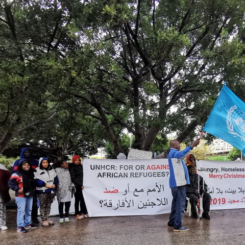 A small group of adults and children stand around a banner that reads, "UNHCR: For or Against African Refugees?" on one side and "Hungry, Homeless, Hopeless: Merry Christmas 2019" on the other. In the center of the image, one man holds the UN Refugee Agency flag. 