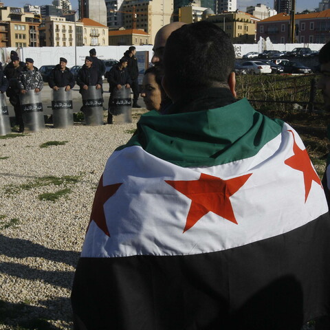 Supporters of the 2011 Syrian uprising, draped in the Syrian opposition flag, gather to commemorate its first anniversary, as members of Lebanon’s Internal Security Forces watch in relaxed shield formation. Martyrs’ Square, Beirut, Lebanon. March 17, 2012. (Marwan Tahtah/The Public Source)