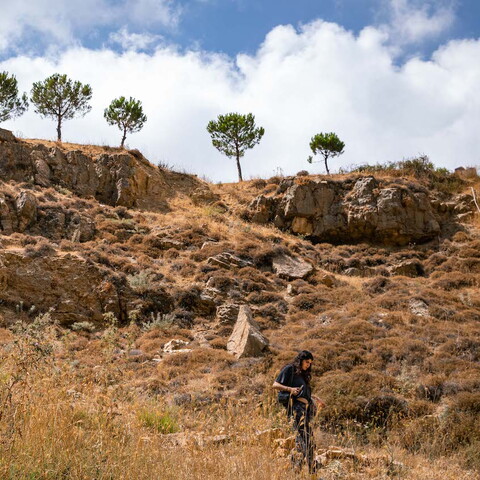 A wide shot showing a young woman dressed in black making her way through a foraging trail down a rocky path. Trees line up the top of the hill and stand tall against a cloudy blue sky.