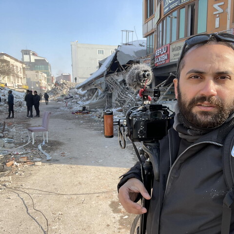 A self-portrait of journalist Issam Abdallah in the foreground, carrying his DSLR camera and microphone, to the background of destroyed buildings. Clear winter day.