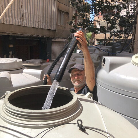 A man in a cap with a white mustache points a thick black hose into a plastic grey water container, and water flows from the hose into the container. There are other such containers around him.