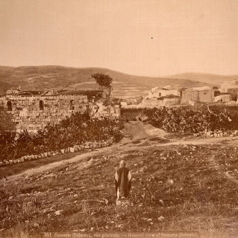 Sepia-toned photograph of ancient stone ruins and surrounding landscape in Samaria (Sebaste), with a person sitting in the foreground. Hills and buildings are visible in the background.