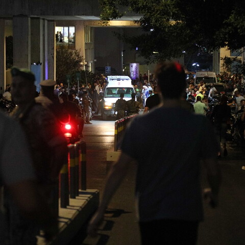 In the foreground are the the backs of men walking toward an ambulance arriving on a narrow street, surrounded by people.