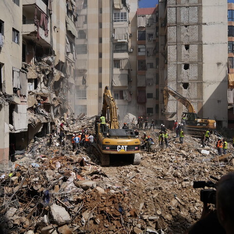 Two bulldozers and several people in high visibility vests stand atop a large mound of rubble, in the middle of a residential area, amid the destruction and ruin of nearby buildings.
