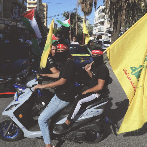 Three male protestors on two motorcycles are carrying the yellow flag of Hezbollah. The Palestinian flag waves in the background.