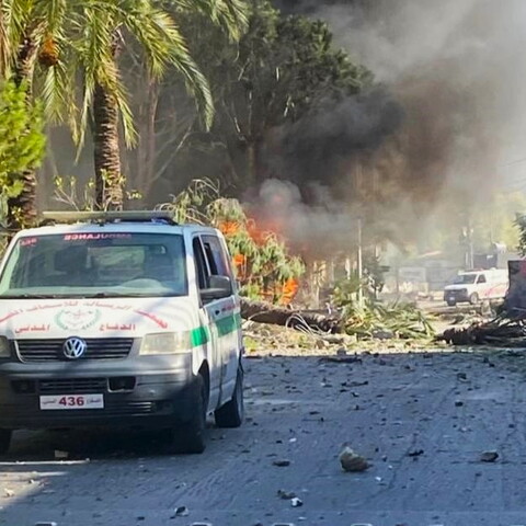 An ambulance on a debris-strewn street with smoke and flames visible in the background. Trees and damaged vehicles are present.